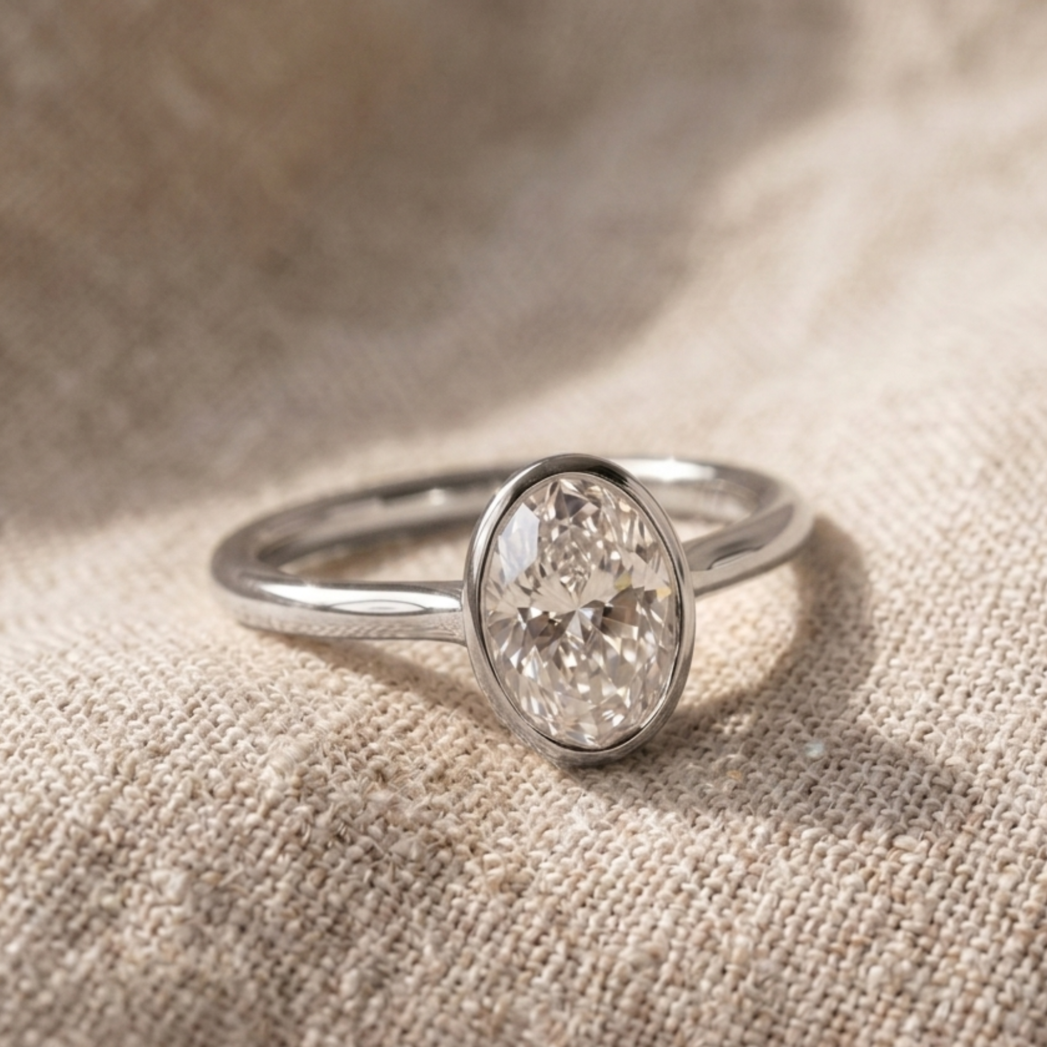A silver ring with an oval-cut clear gemstone in a bezel setting, resting on a textured beige linen fabric.