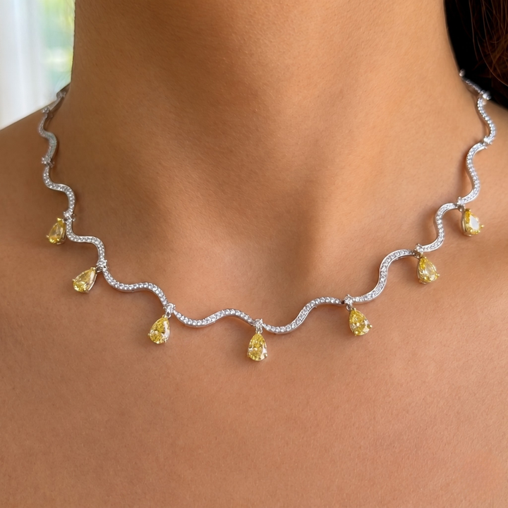 A close-up of a model's neck and upper chest wearing a wavy silver-toned necklace adorned with small clear stones and five yellow teardrop gemstones.