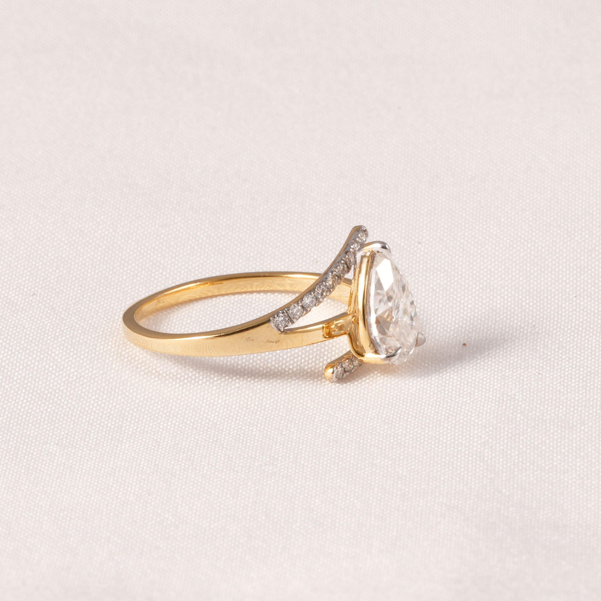A gold ring with a pear-cut clear center stone and small pavé-set clear accent stones on a curved band, photographed on a light neutral fabric background.