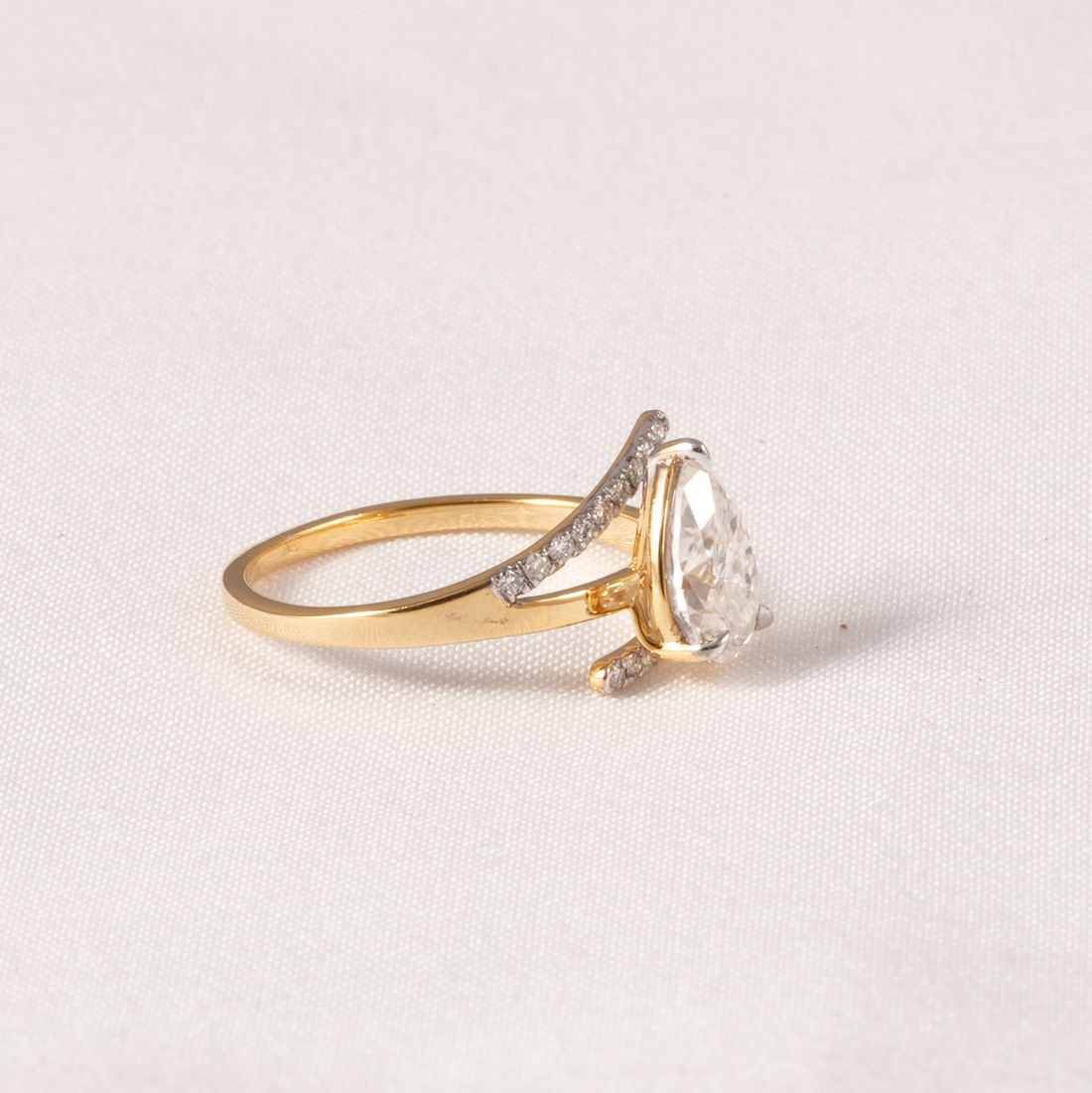 A gold ring with a pear-cut clear center stone and small pavé-set clear accent stones on a curved band, photographed on a light neutral fabric background.