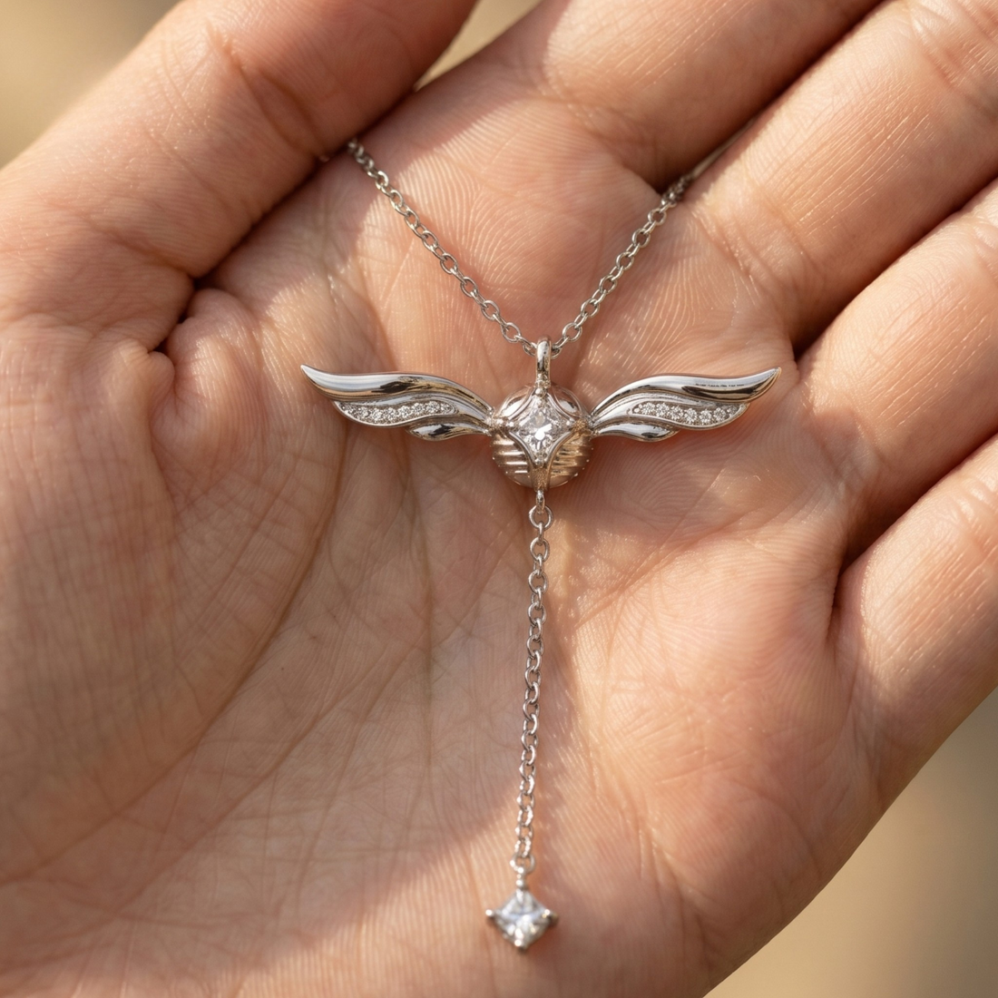 A silver-toned winged pendant necklace with a dangling chain and clear gemstones, held in the palm of a hand.