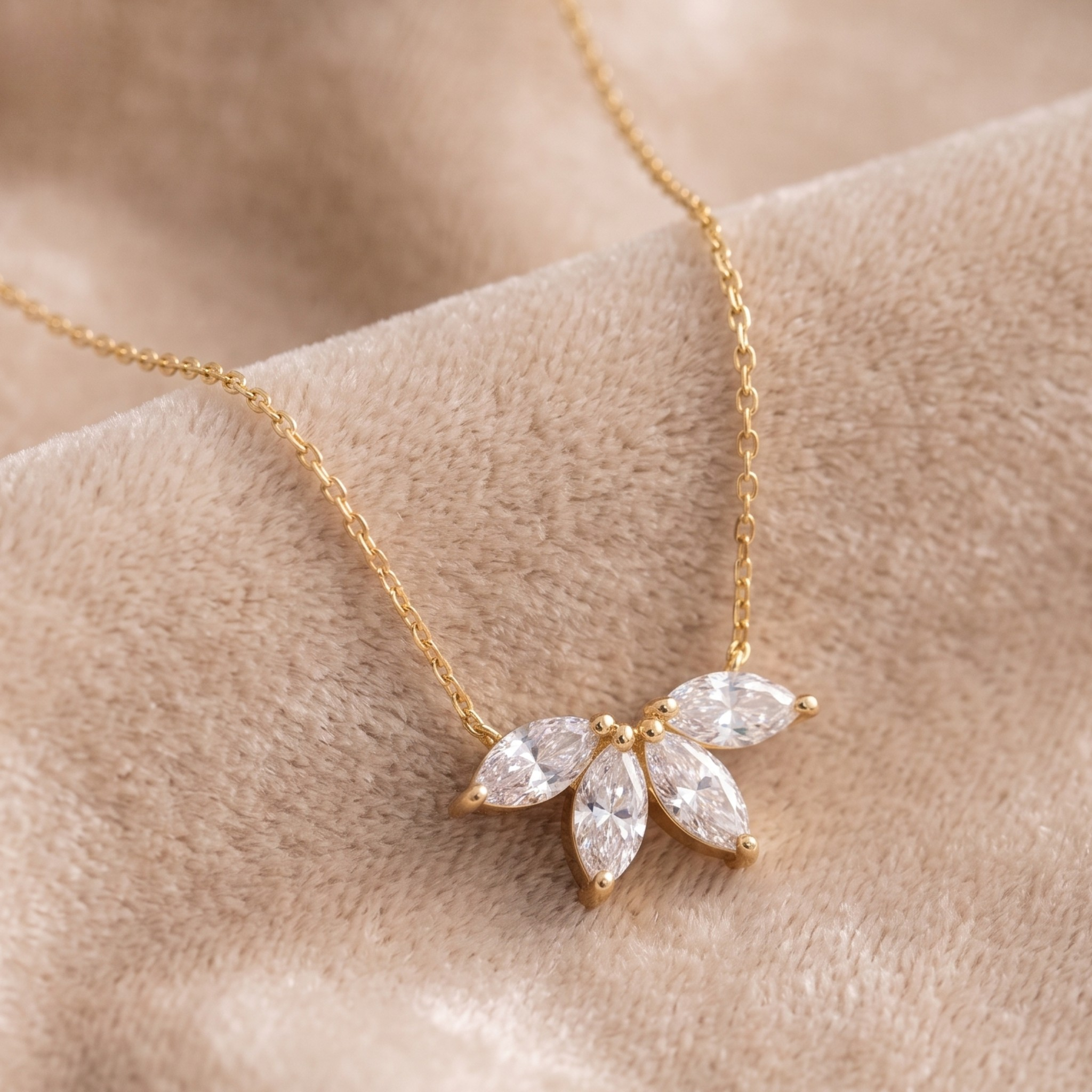 A delicate gold chain necklace with a small flower-like pendant made of clear marquise-cut stones, displayed on a soft beige fabric surface.