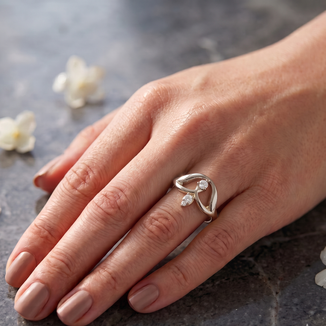 A silver ring with a small clear gemstone, worn on a woman's hand resting on a dark textured surface with white flowers in the background.