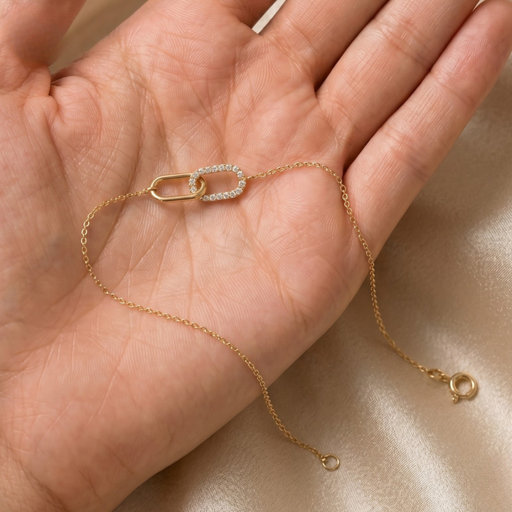 A delicate gold chain bracelet with a double-link design, one side featuring small clear stones, resting on an open hand over a beige satin fabric background.