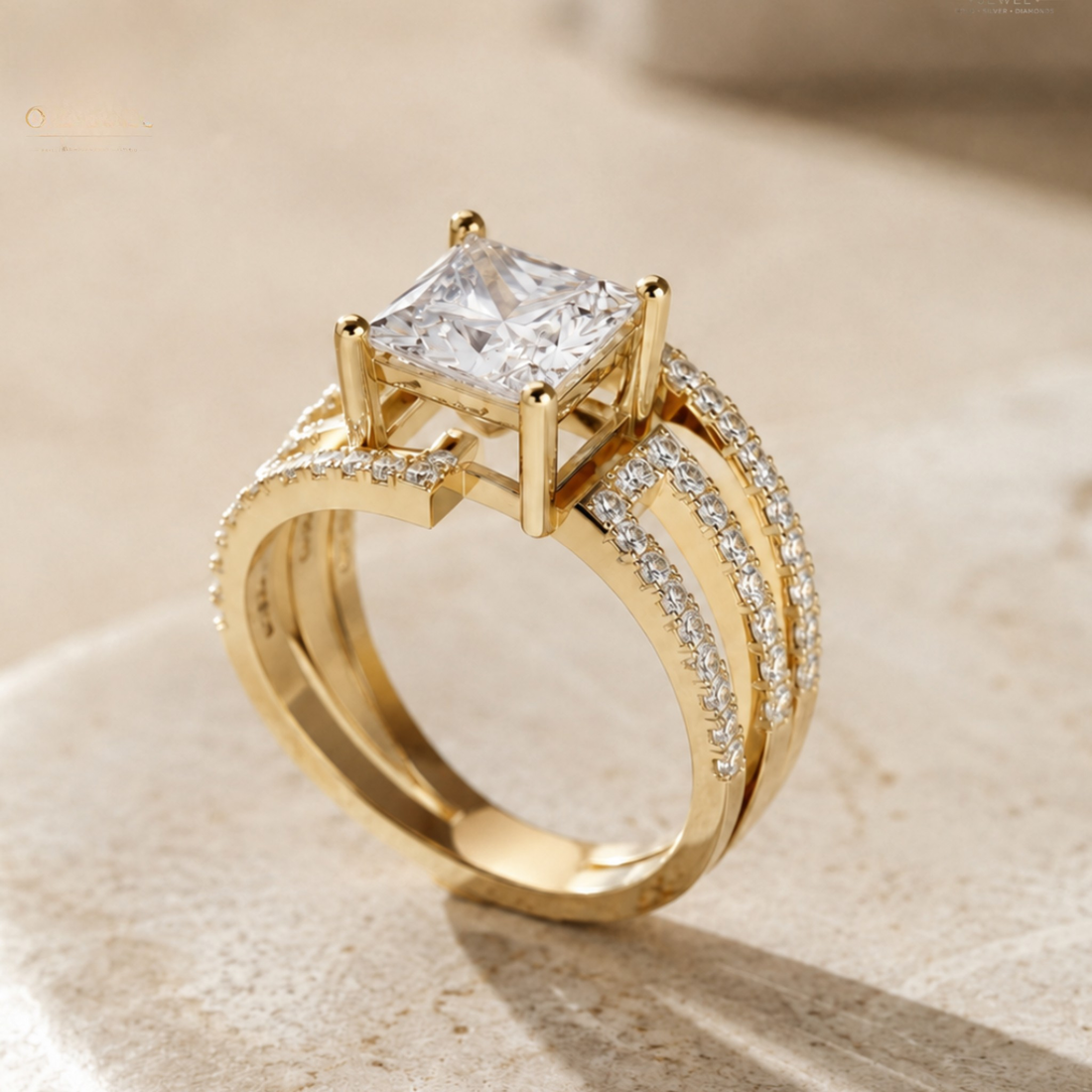 A gold ring featuring a princess-cut clear center stone and pavé-set accent stones along curved bands, photographed on a soft beige stone surface.