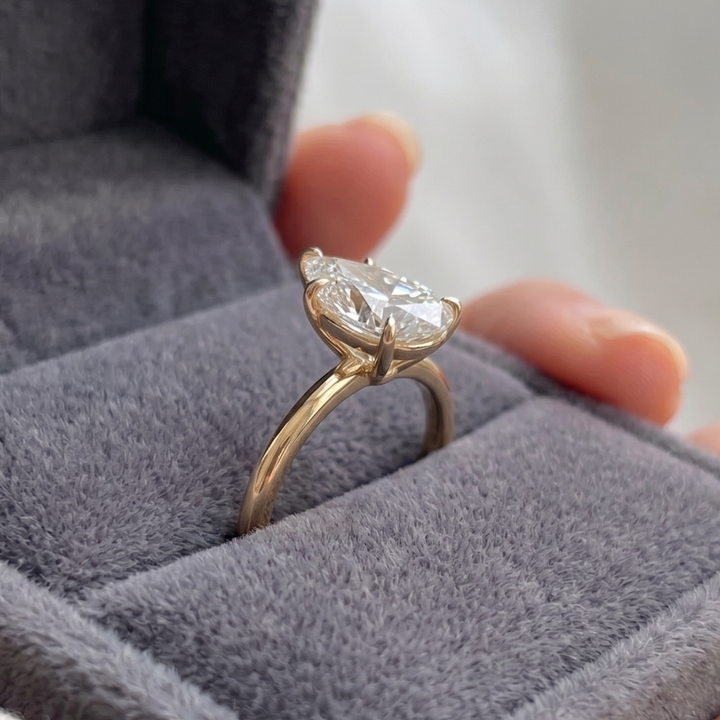 A gold ring with a clear gemstone resting on a gray velvet ring box, with fingers holding the box in the background.