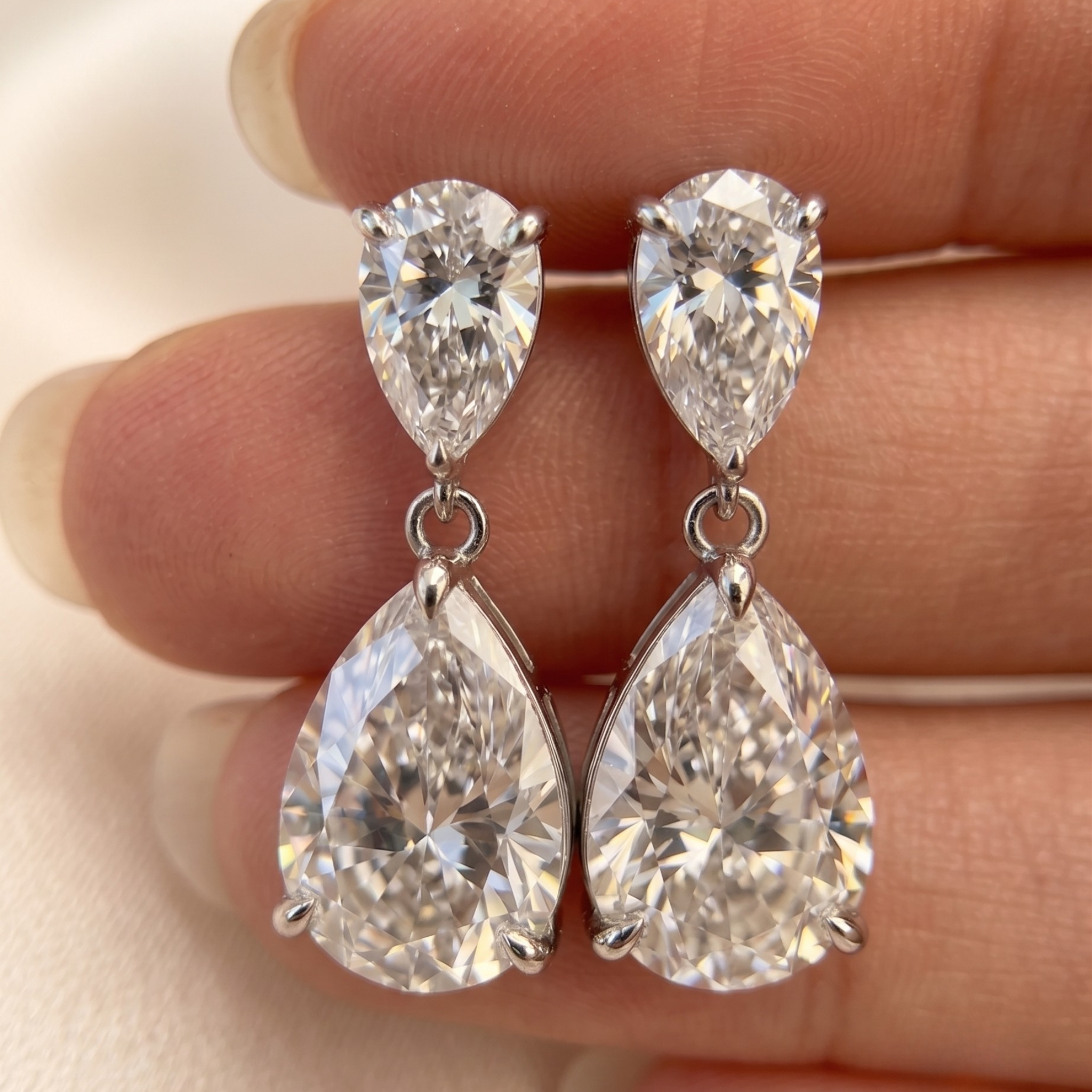 A pair of silver-tone drop earrings with pear-cut clear gemstones, held between fingers against a soft light background.