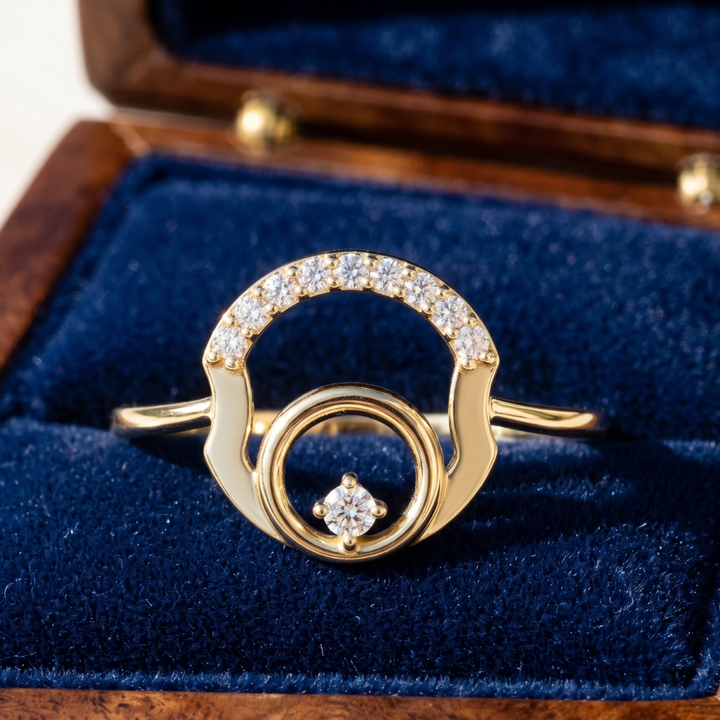 A gold ring with a double-ring design, a curved top set with small clear stones, and a single central clear stone, displayed on a blue velvet ring box.