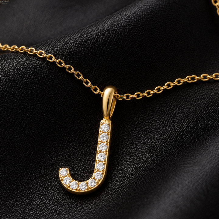 A gold initial pendant necklace featuring the letter 'J' encrusted with small clear stones, displayed on a textured black fabric background.