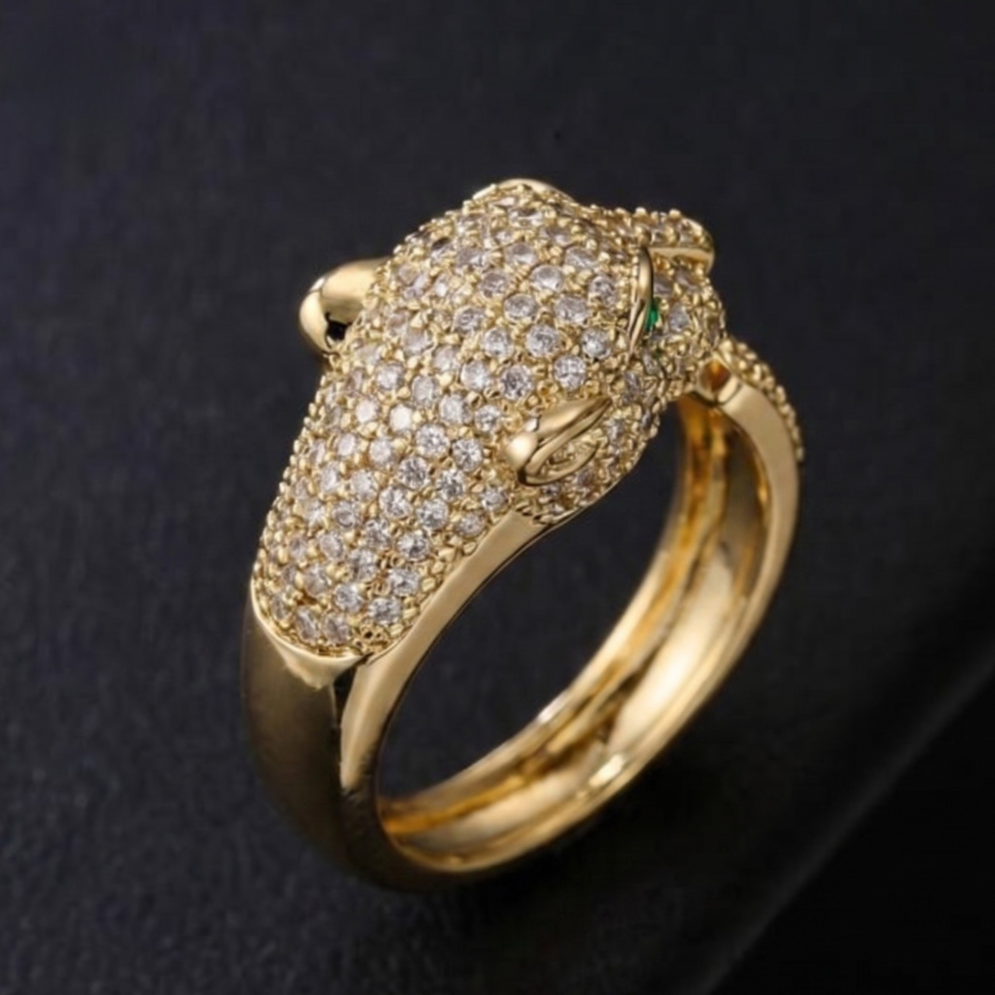 A gold ring featuring a detailed animal head motif, heavily set with small clear pavé stones and accented with a tiny green stone, shown on a dark textured background.