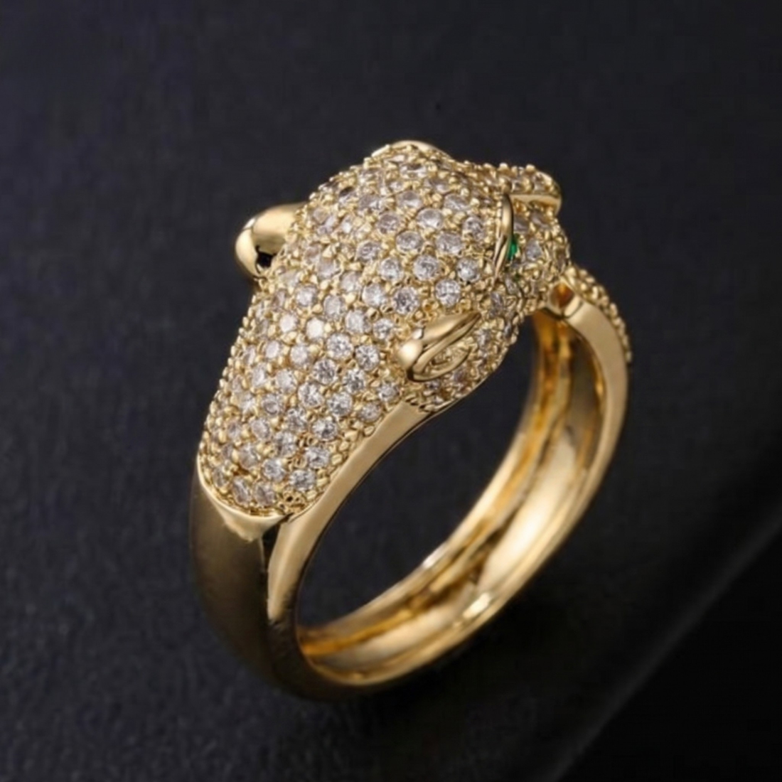A gold ring featuring a detailed animal head motif, heavily set with small clear pavé stones and accented with a tiny green stone, shown on a dark textured background.