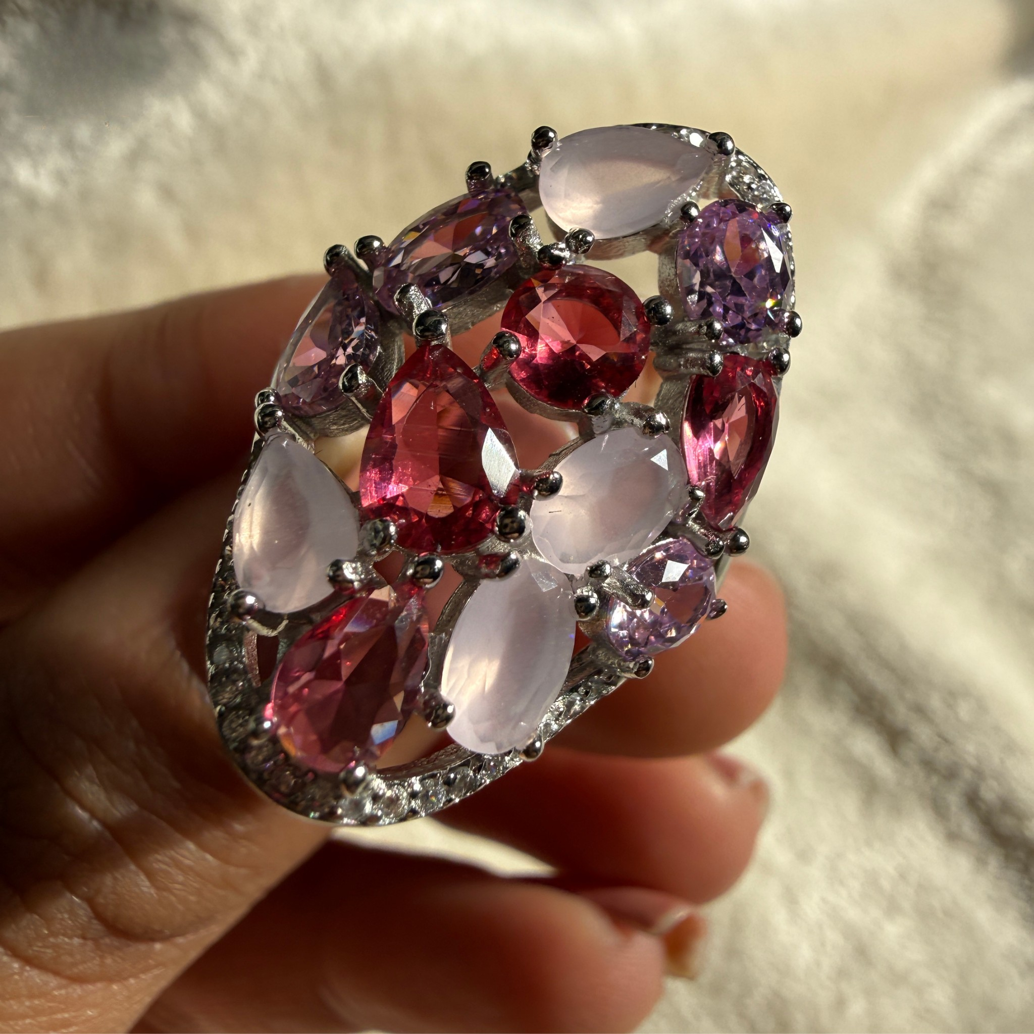 A large statement ring held in a hand, featuring oval and marquise-cut pink and purple gemstones in a silver-toned setting with small clear accent stones, against a soft white background.