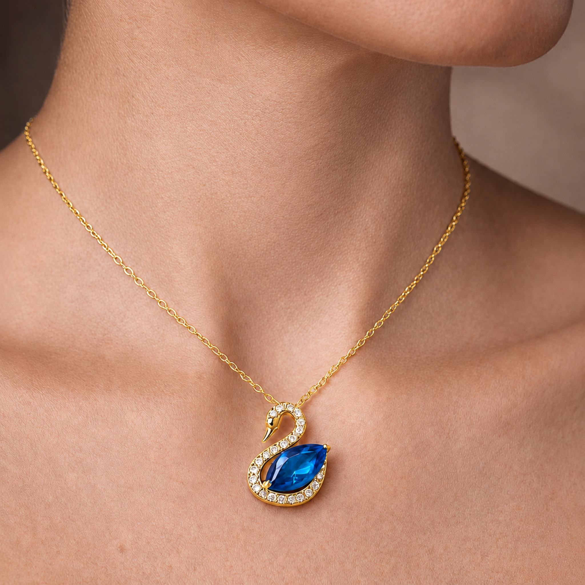 A gold necklace with a swan pendant featuring a large blue teardrop gemstone and small clear stones, shown worn on a model's neck.