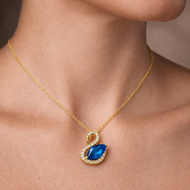 A gold necklace with a swan pendant featuring a large blue teardrop gemstone and small clear stones, shown worn on a model's neck.