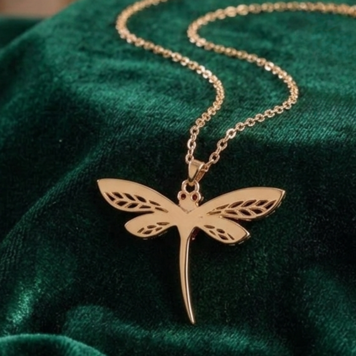 A gold dragonfly pendant necklace on a delicate gold chain, displayed on a dark green velvet fabric background.