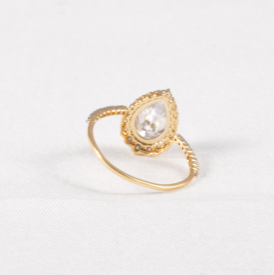 A gold ring featuring a teardrop-shaped clear gemstone in a decorative bezel setting, with a twisted rope-style band, shown on a white textured background.