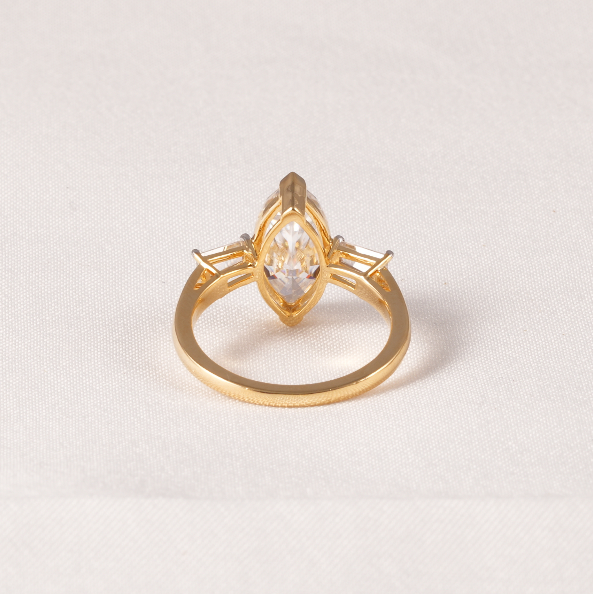 A gold ring featuring a marquise-cut clear gemstone in a prong setting, with smaller side stones, shown on a textured off-white fabric background.