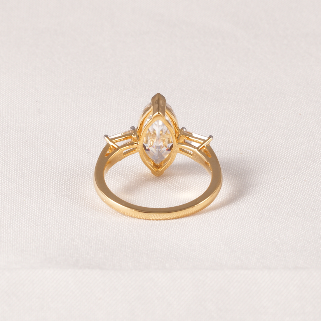A gold ring featuring a marquise-cut clear gemstone in a prong setting, with smaller side stones, shown on a textured off-white fabric background.