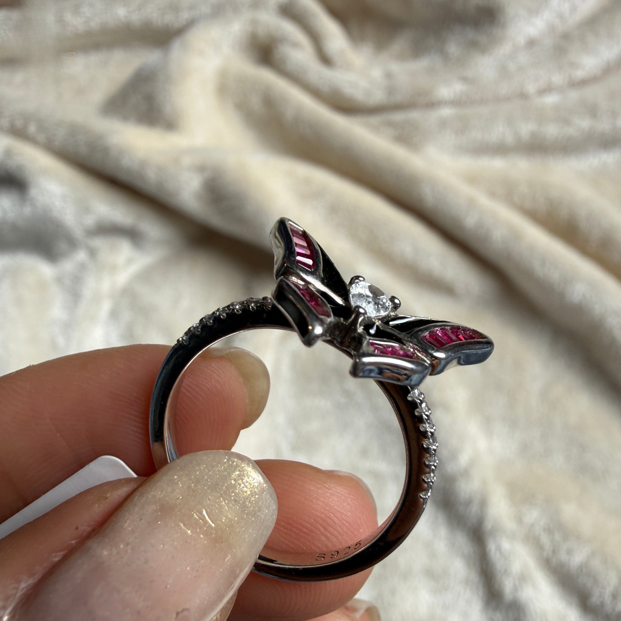 A close-up of a hand holding a silver ring with a butterfly design, featuring pink gemstone accents and a clear central stone, set against a soft beige fabric background.