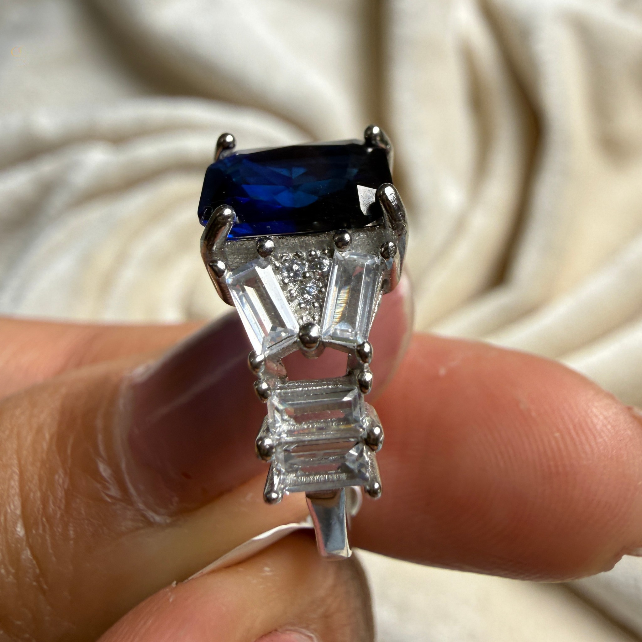 A silver ring with a rectangular dark blue sapphire center stone and baguette-cut clear stones, held between fingers against a draped cream fabric background.