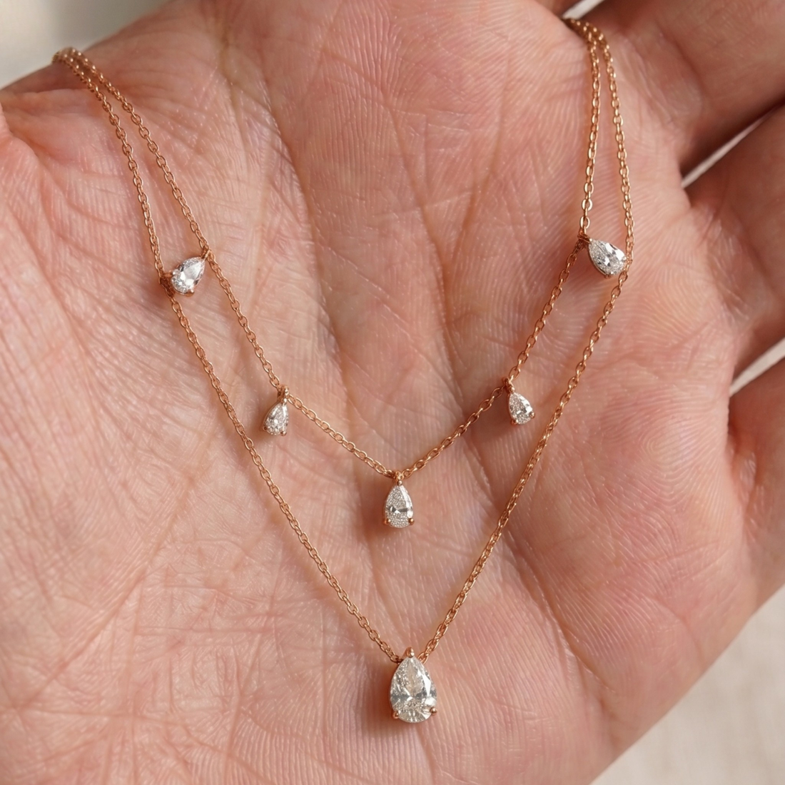 A delicate rose gold double-strand necklace with small teardrop crystal charms and a larger teardrop pendant, displayed on an open hand.