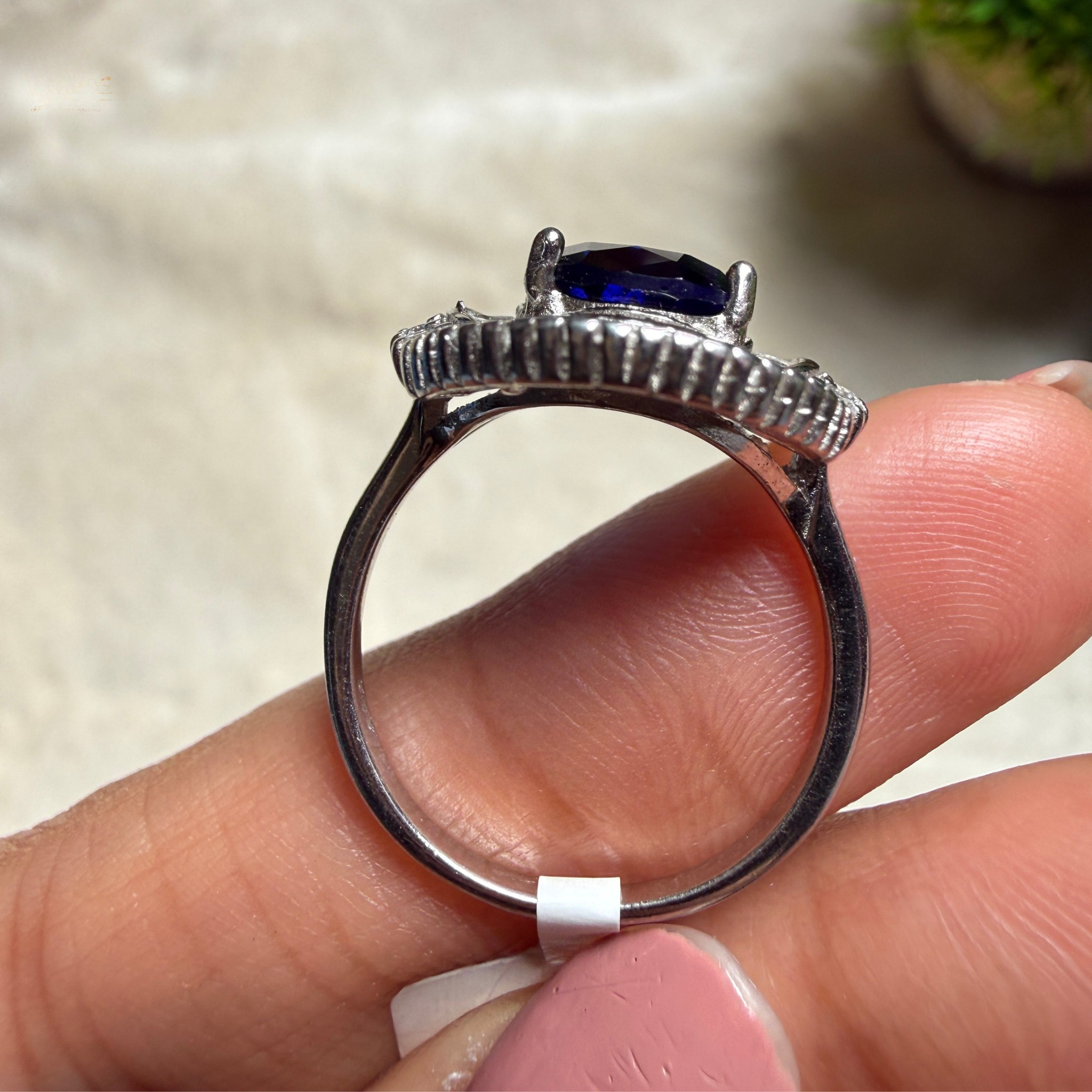 A silver ring with a deep blue rectangular gemstone, held on a person's fingers against a soft beige background.