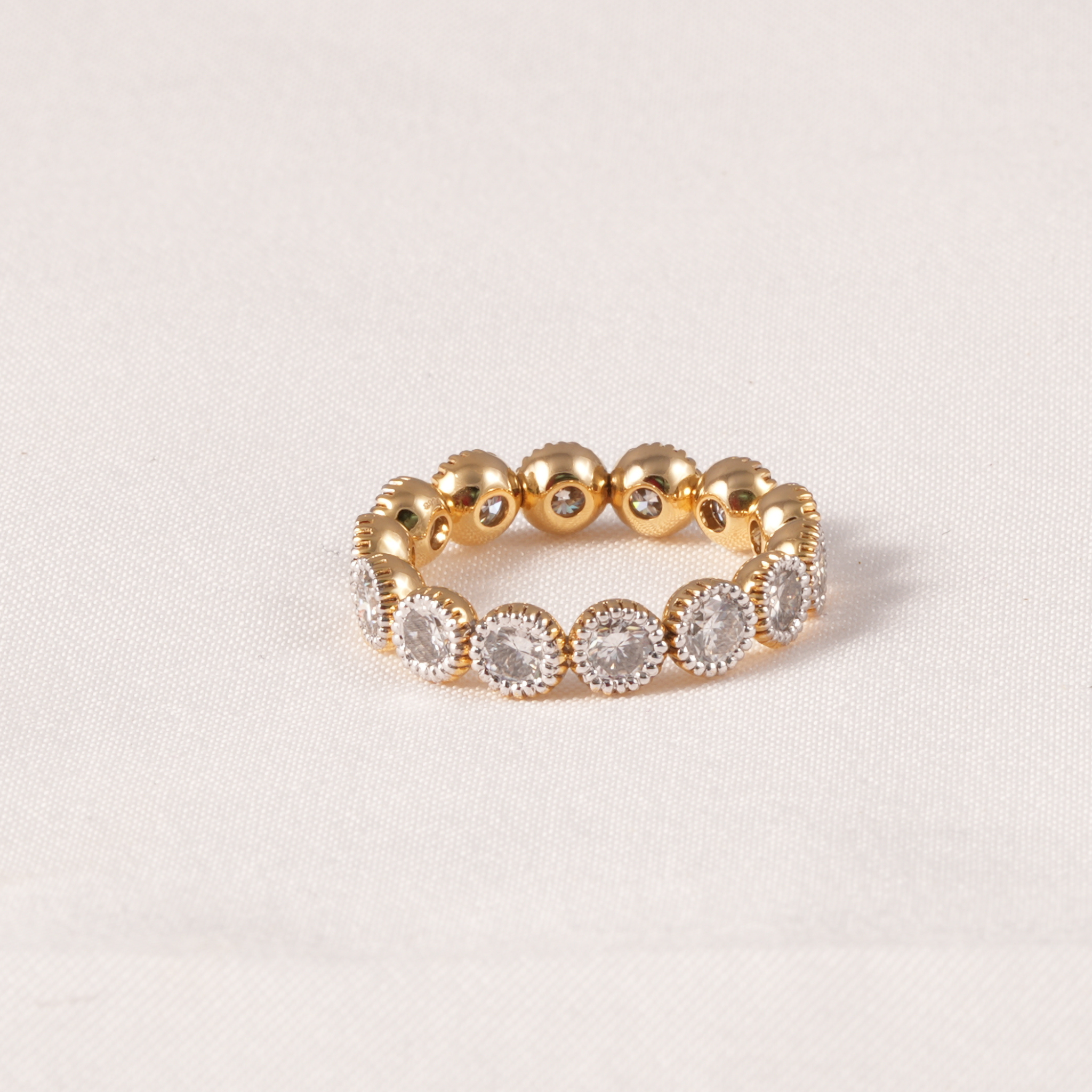 A gold eternity-style band ring featuring a row of round clear gemstones in halo settings, photographed on a light beige fabric background.