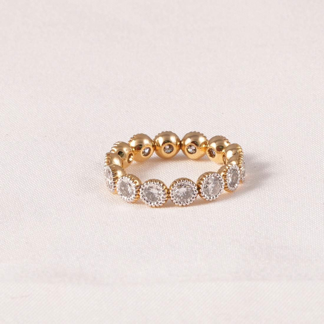 A gold eternity-style band ring featuring a row of round clear gemstones in halo settings, photographed on a light beige fabric background.