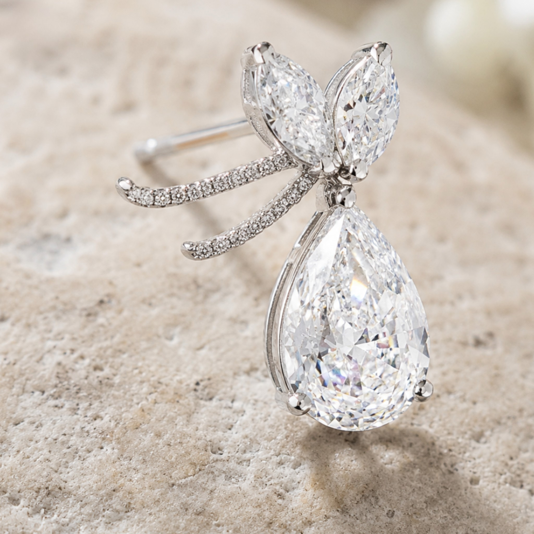 A silver butterfly ring with a large pear-cut clear gemstone and smaller marquise and round-cut stones, displayed on a textured beige stone surface.