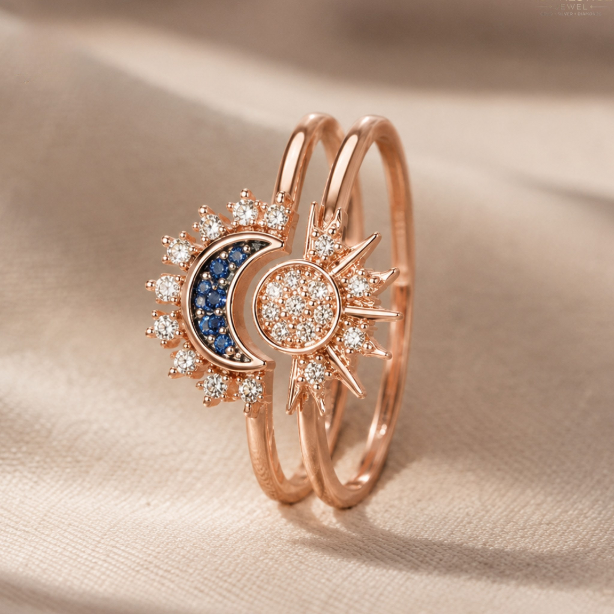 A rose gold double-band ring featuring a moon and sun design, embellished with clear pavé stones and a dark blue crescent moon center, displayed on a beige fabric background.