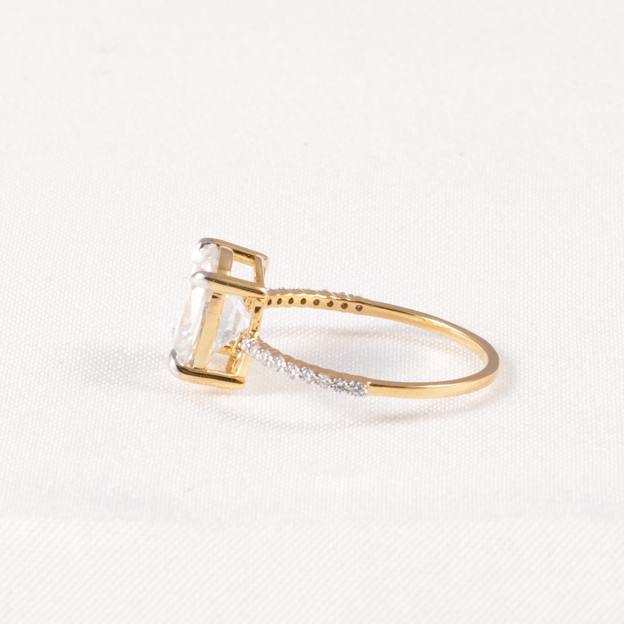 A yellow gold ring featuring a large rectangular clear gemstone in a prong setting, with a pavé band accented by small clear stones, shown on a textured white background.