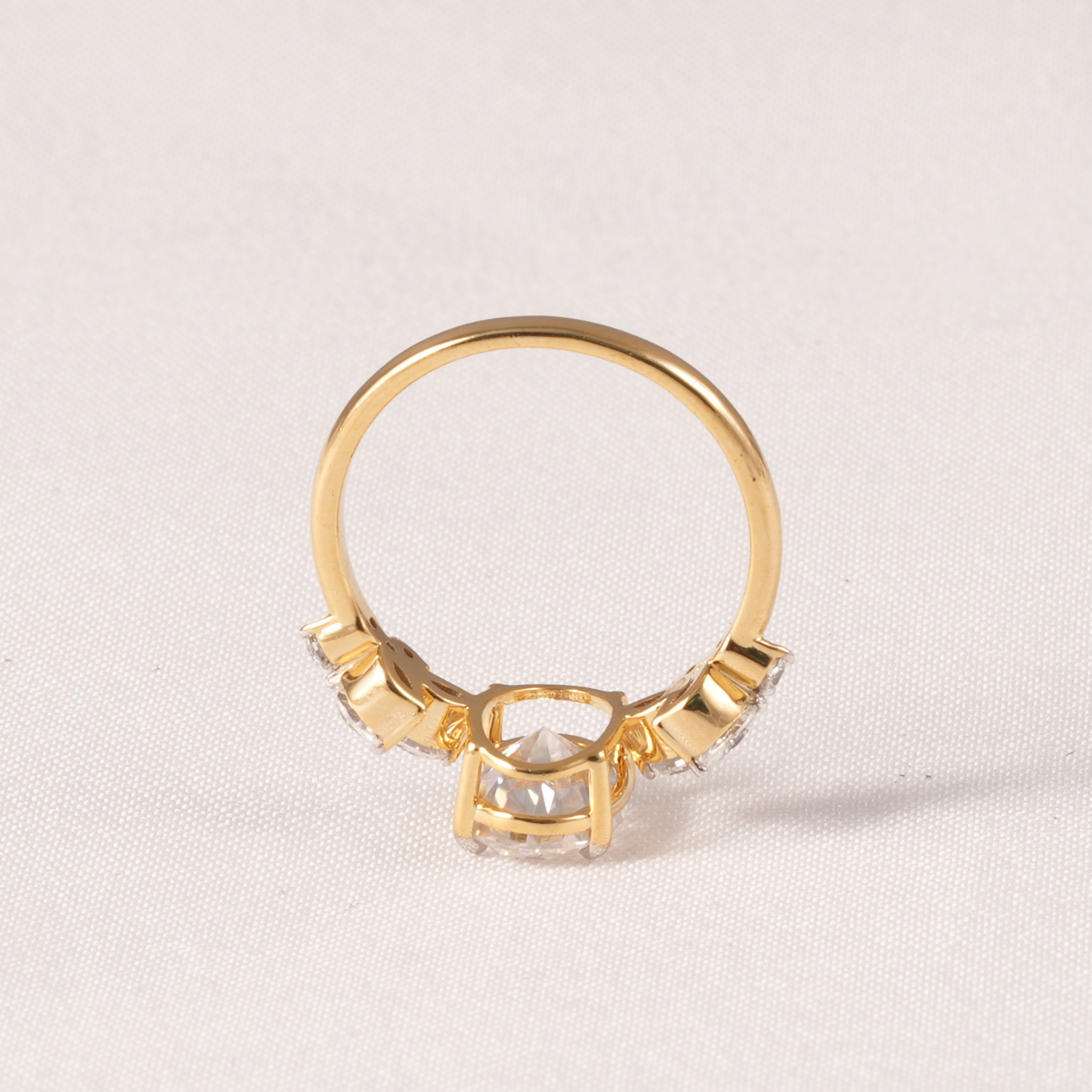 A gold ring with a central clear rectangular gemstone and smaller clear stones on either side, photographed on a light beige textured fabric background.