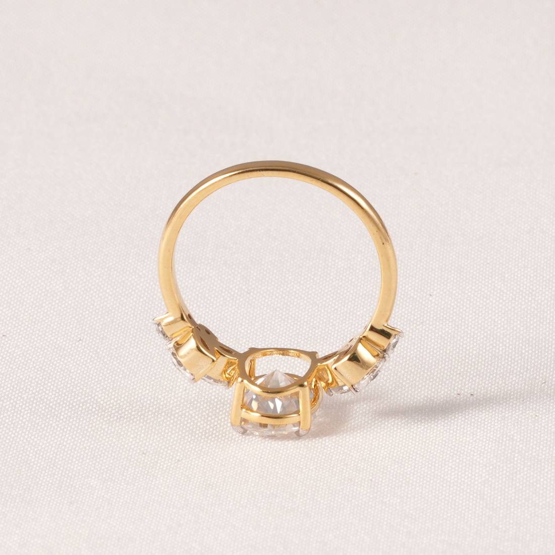 A gold ring with a central clear rectangular gemstone and smaller clear stones on either side, photographed on a light beige textured fabric background.