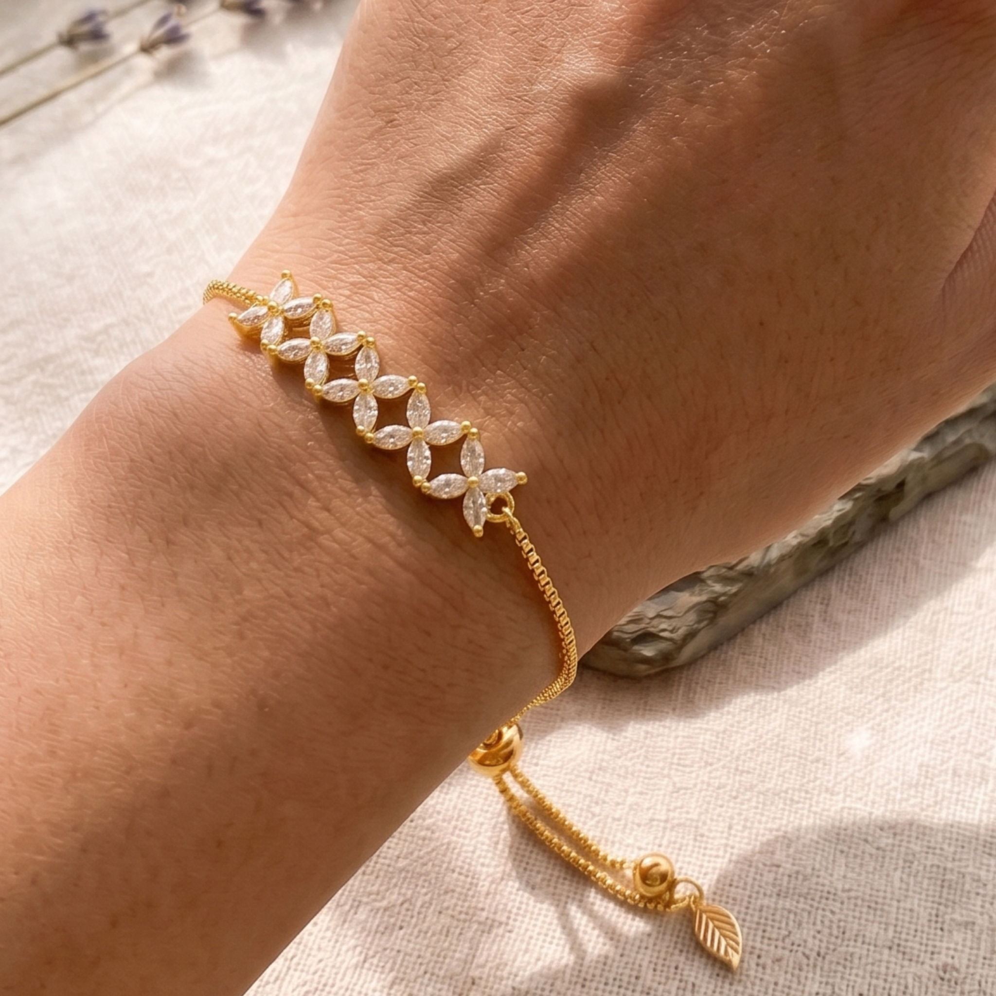 A gold bracelet with a row of clear crystal flower motifs worn on a person's wrist, resting on a textured light beige fabric surface.