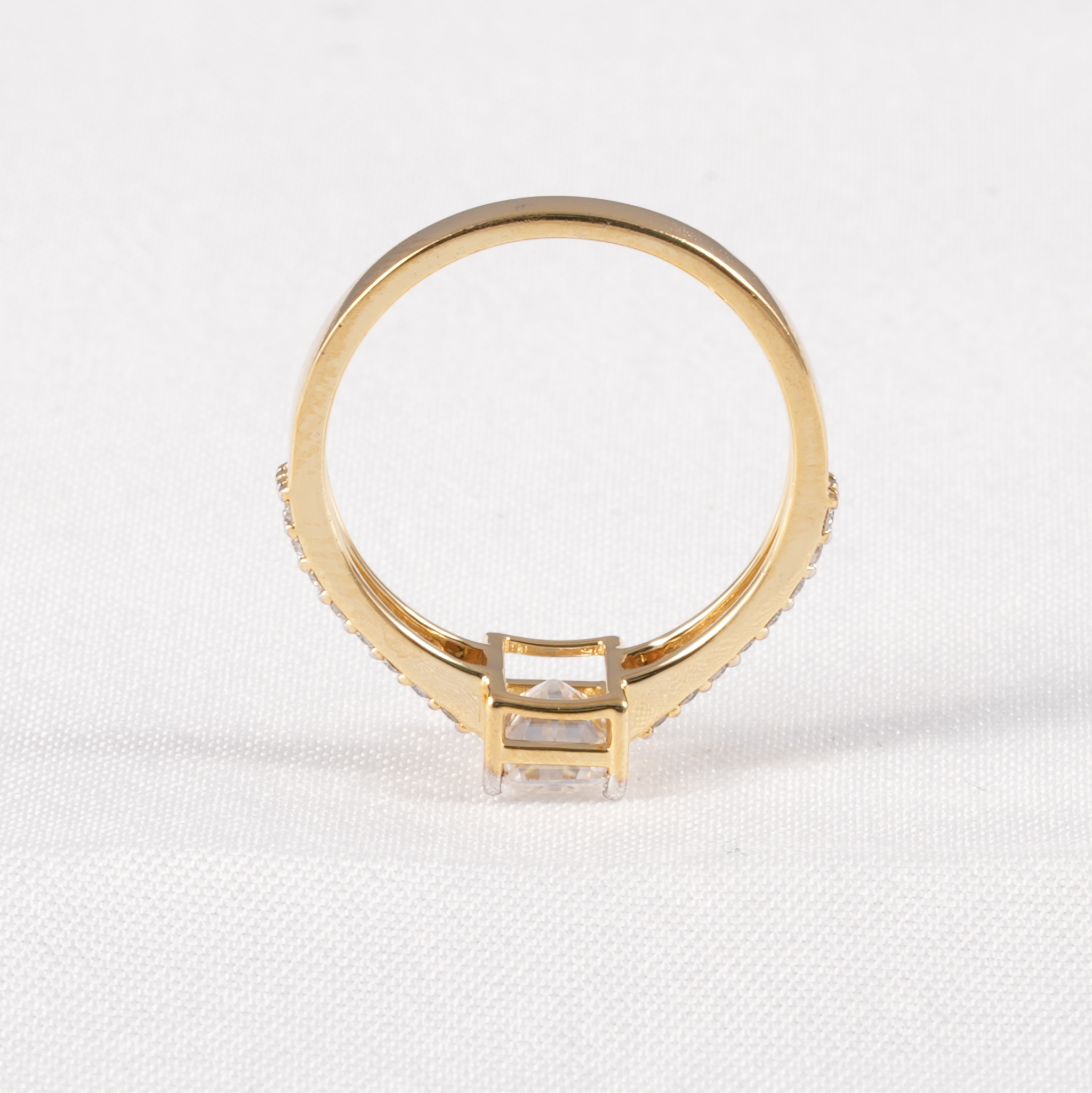 A yellow gold ring with a square-cut clear center stone and small clear stones along the shoulders, photographed on a white textured fabric background.