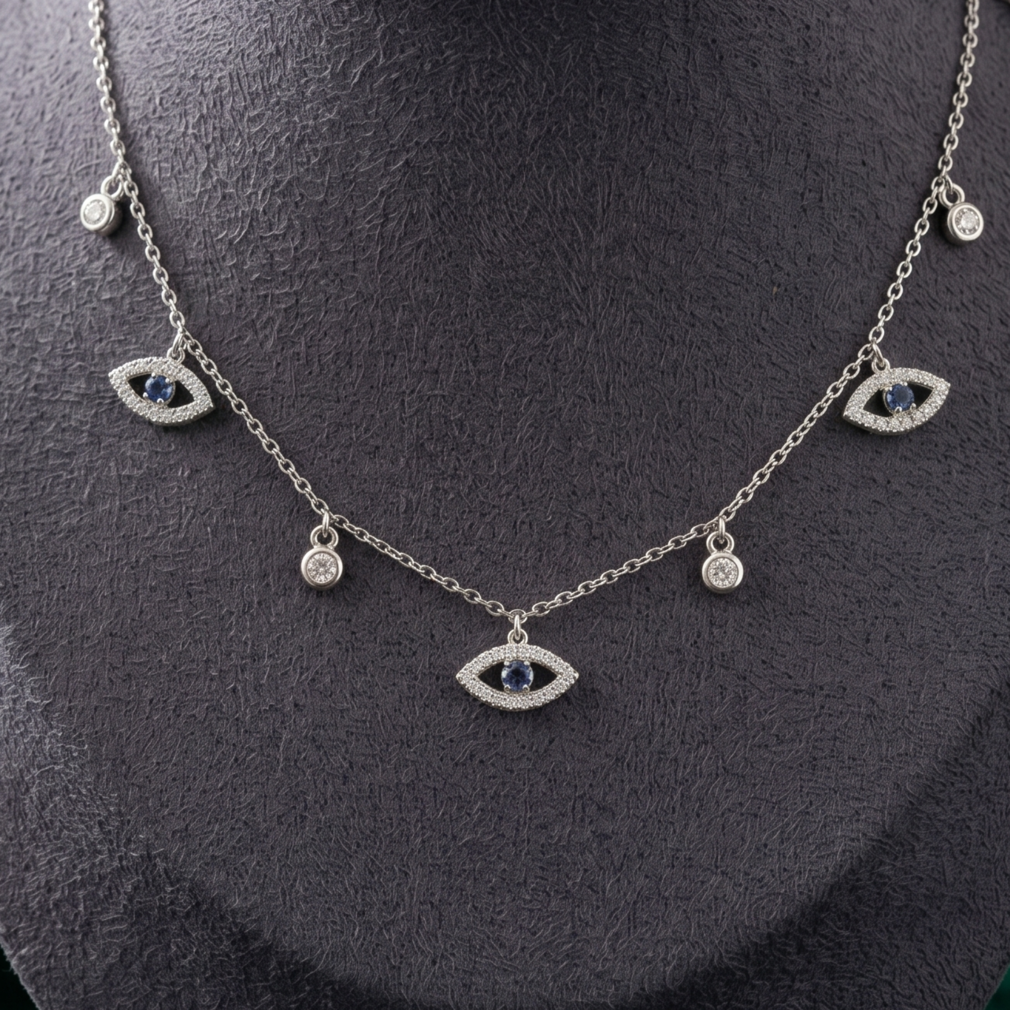 A silver-tone evil eye necklace with dark blue gemstone centers and clear crystal accents, displayed on a textured black jewelry bust.