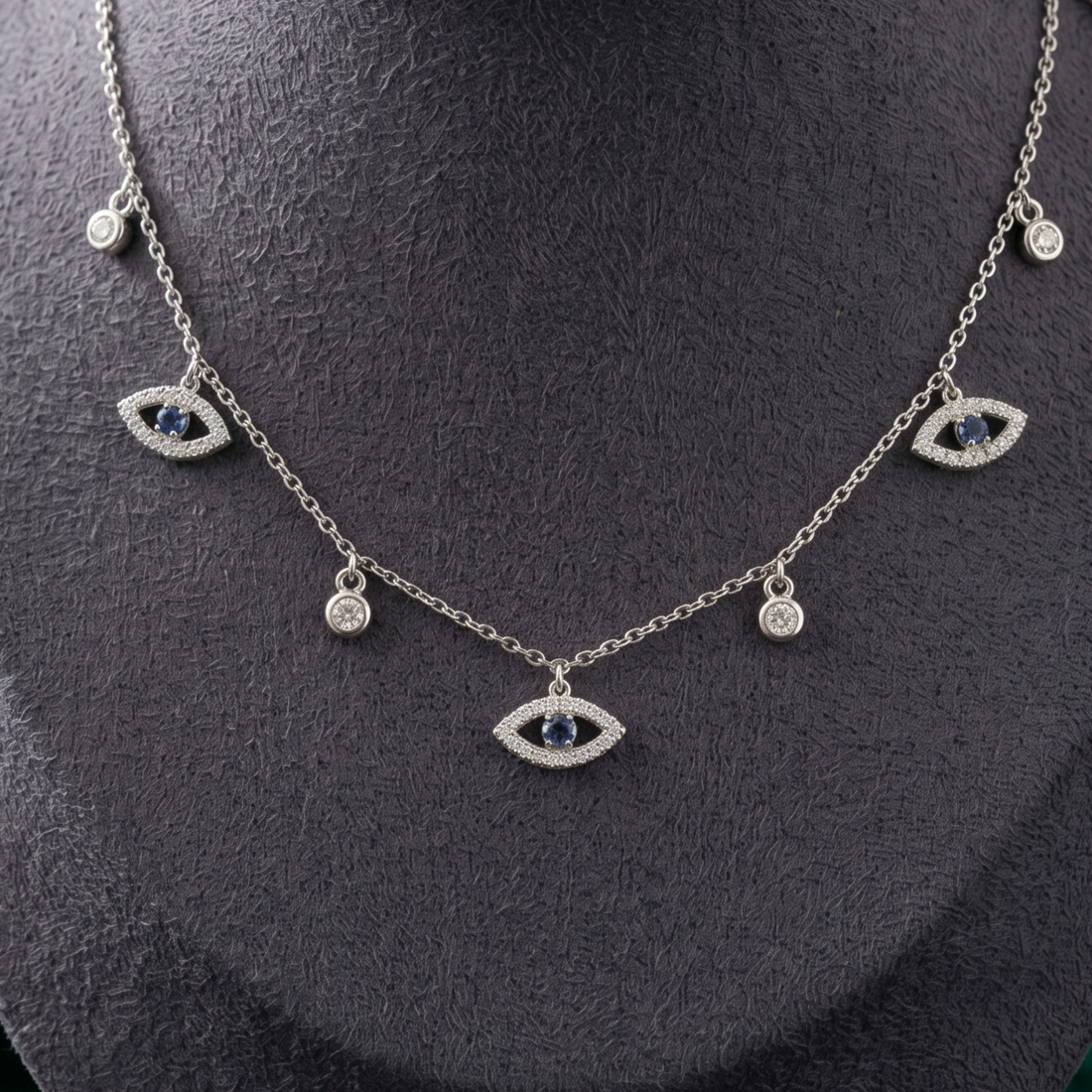 A silver-tone evil eye necklace with dark blue gemstone centers and clear crystal accents, displayed on a textured black jewelry bust.