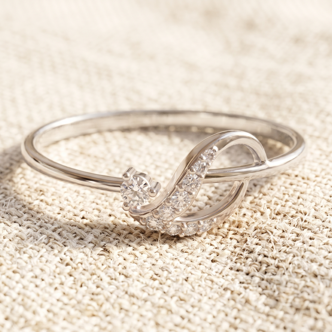 A silver ring with a stylized intertwined loop design, accented by a round clear gemstone and a curved row of small clear stones, displayed on a textured beige fabric background.