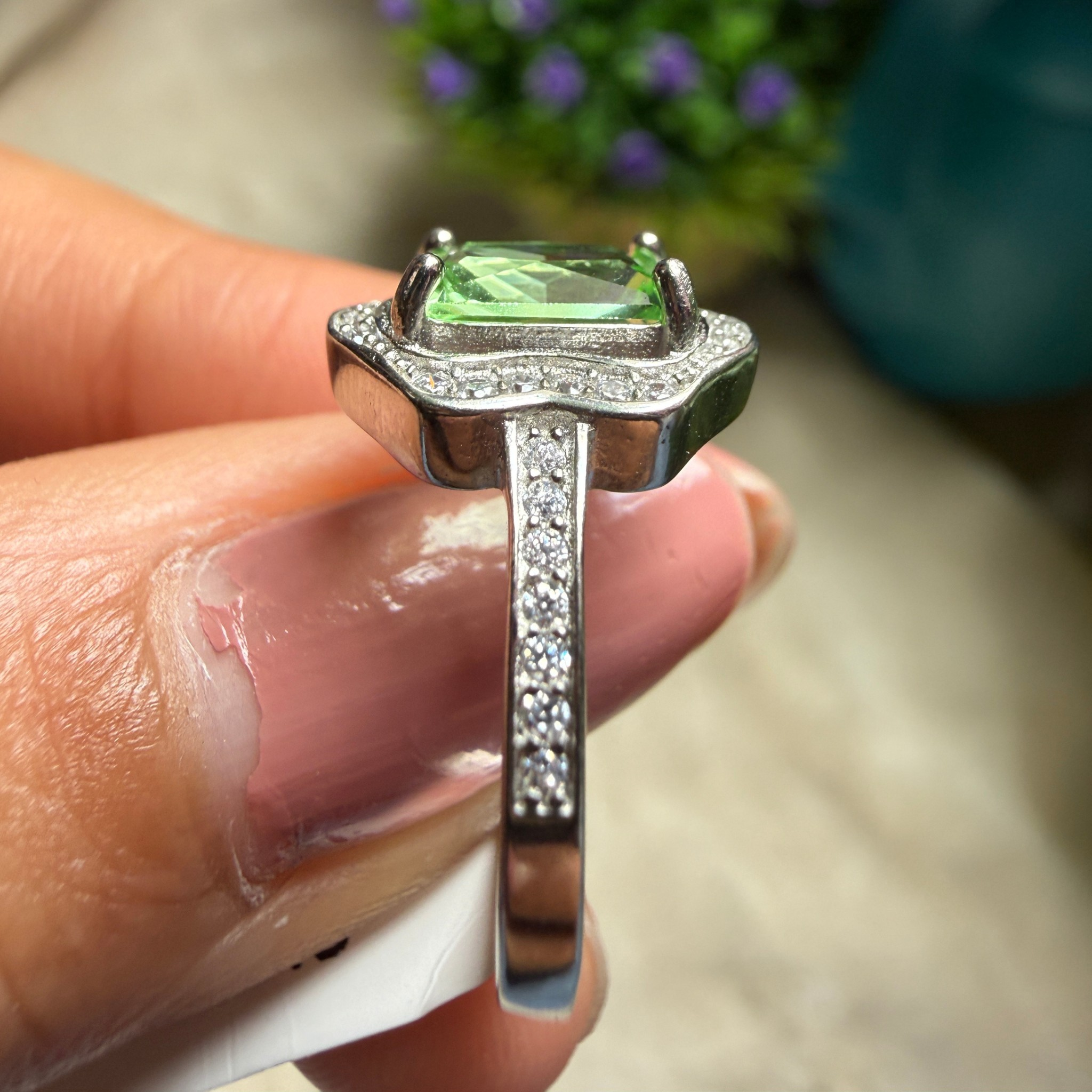 A close-up of a hand holding a silver ring with a green rectangular gemstone and small clear accent stones.