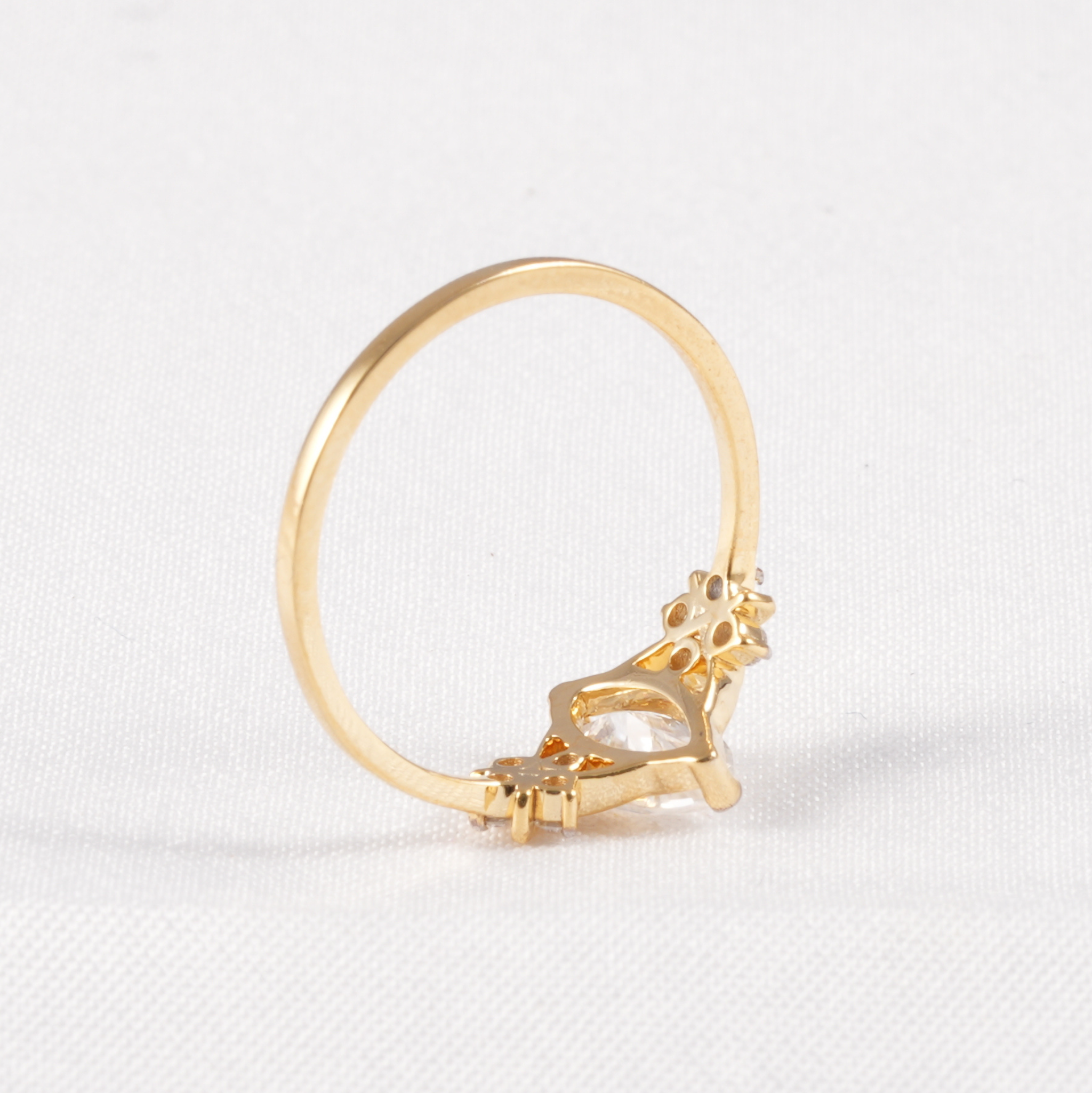 A gold ring with a decorative floral crown-like setting and a clear marquise-cut gemstone, photographed on a white textured fabric background.