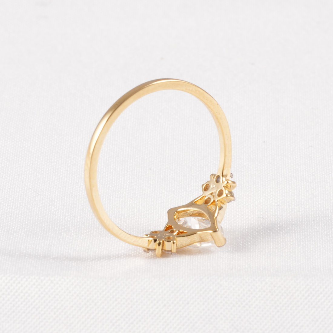 A gold ring with a decorative floral crown-like setting and a clear marquise-cut gemstone, photographed on a white textured fabric background.