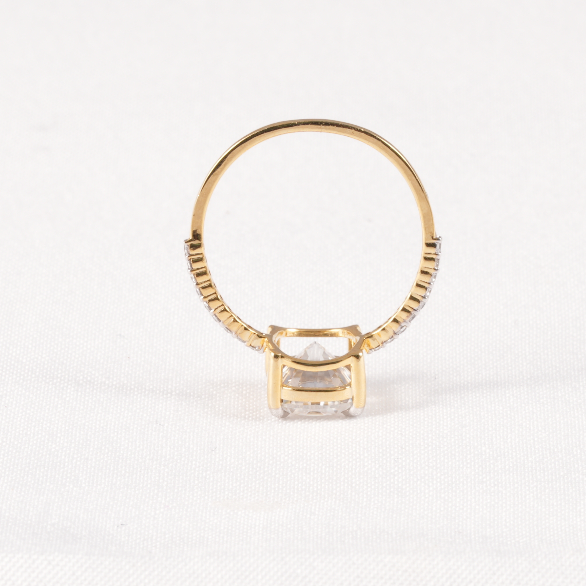 A yellow gold ring with a square-cut clear gemstone in a prong setting, accented with small pavé diamonds along the band, shown on a white textured background.