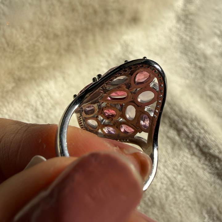 A close-up of a silver-toned ring with multiple oval pink gemstones, held between fingers against a soft beige textured background.