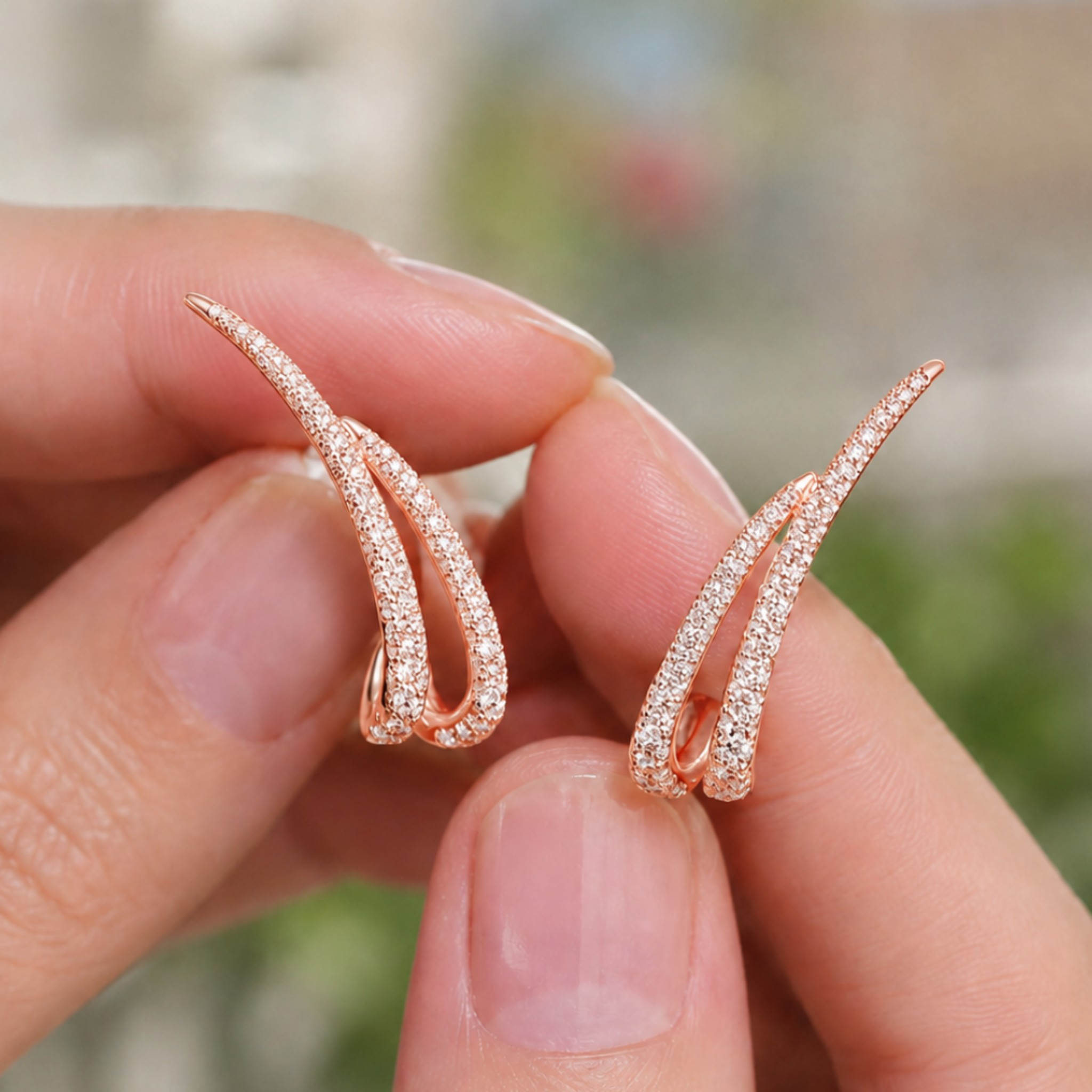 A pair of rose gold huggie hoop earrings paved with small clear stones, held between two fingers against a blurred outdoor background.