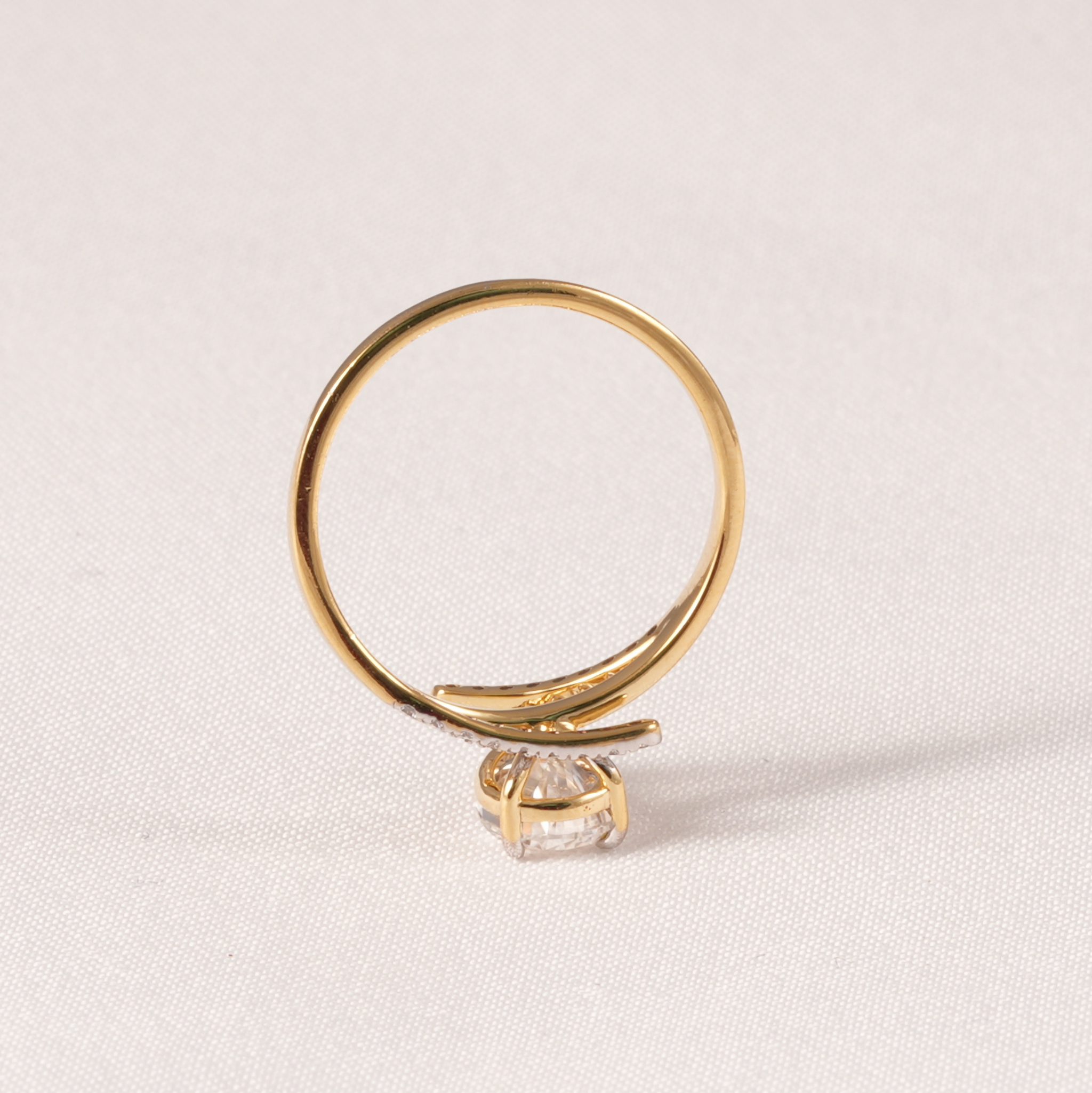 A gold ring with a curved band and a single clear gemstone, photographed on a light beige textured fabric background.