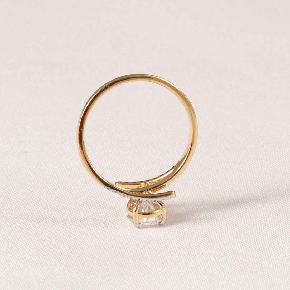 A gold ring with a curved band and a single clear gemstone, photographed on a light beige textured fabric background.