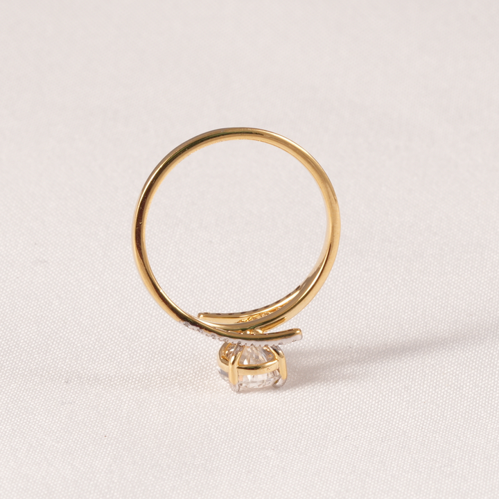 A gold ring with a curved band and a single clear gemstone, photographed on a light beige textured fabric background.