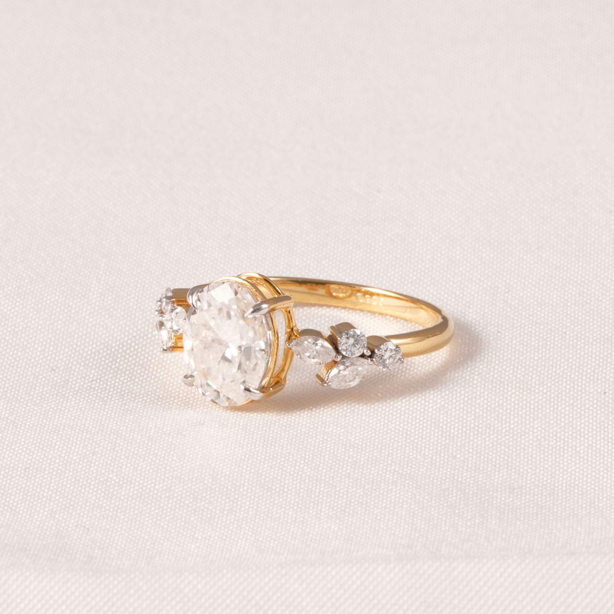 A yellow gold ring featuring a large round-cut clear center stone and smaller marquise-cut side stones, photographed on a light beige textured fabric background.