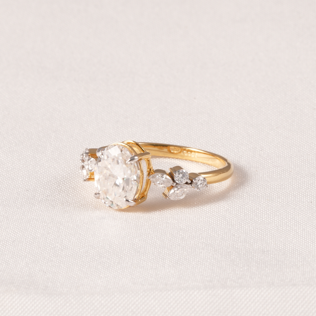 A yellow gold ring featuring a large round-cut clear center stone and smaller marquise-cut side stones, photographed on a light beige textured fabric background.