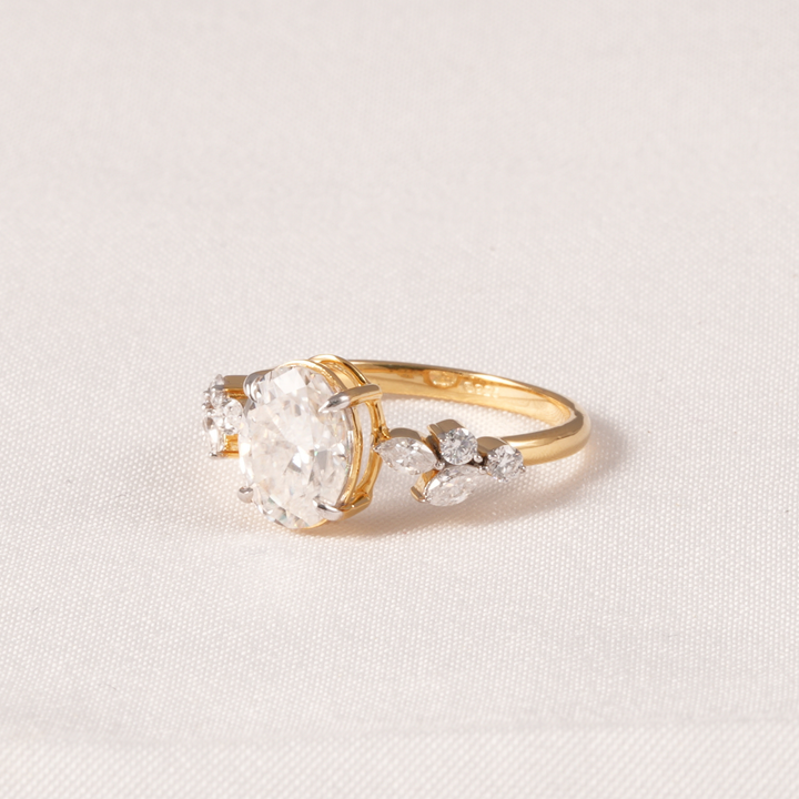 A yellow gold ring featuring a large round-cut clear center stone and smaller marquise-cut side stones, photographed on a light beige textured fabric background.